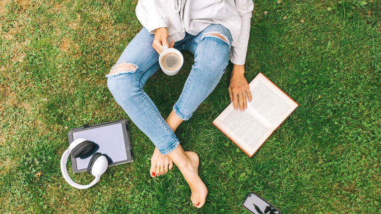 Young woman sitting on the grass drinking coffee and reading a book enjoys outdoor recreation.