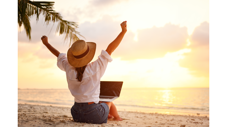 Rear view of young female freelancer in straw hat celebrating success. while working remotely on the tropical sandy beach at sunset