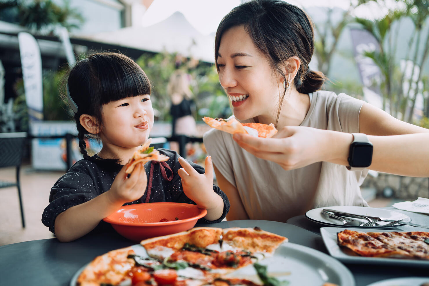 Joyful young Asian mother and lovely little daughter enjoying pizza lunch in an outdoor restaurant. Family enjoying bonding time and a happy meal together. Family and eating out lifestyle