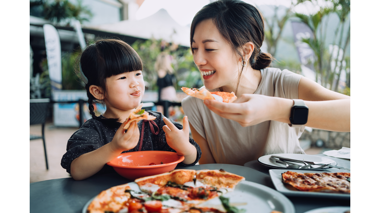 Joyful young Asian mother and lovely little daughter enjoying pizza lunch in an outdoor restaurant. Family enjoying bonding time and a happy meal together. Family and eating out lifestyle