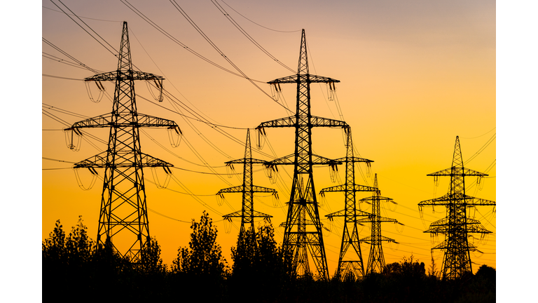 Power pylons reache into the sunset sky. Silhouettes of big trees under energy transmission towers.