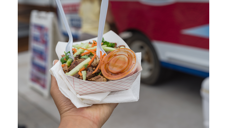 To go take away paper food tray of chicken teriyaki with chili sauce in deli containers, napkins, fork and spoon with blurry food truck background