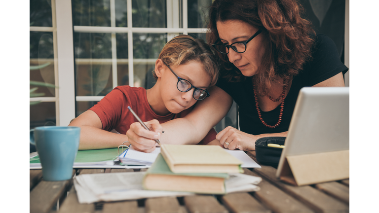 Young student doing homework at home with school books, newspaper and digital pad helped by his mother. Mum writing on the copybook teaching his son. Education, family lifestyle, homeschooling concept