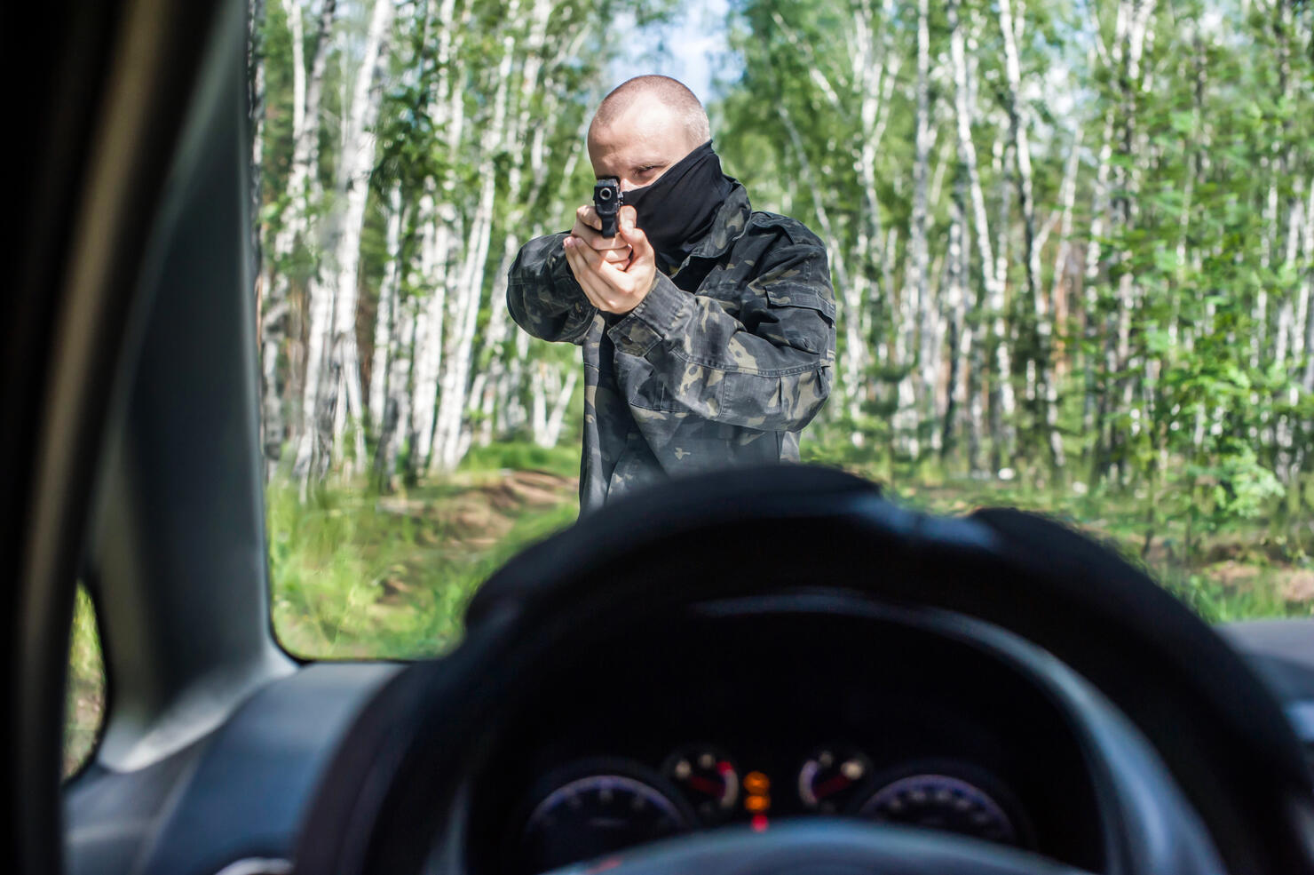 a man in camouflage with a pistol aims at the driver of a car on the road in the forest.