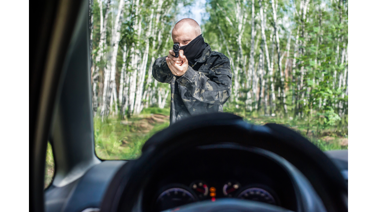 a man in camouflage with a pistol aims at the driver of a car on the road in the forest.