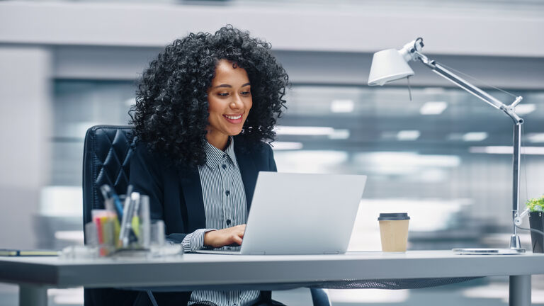 Modern Office: Black Businesswoman Sitting at Her Desk Working on a Laptop Computer. Smiling Successful African American Woman working with Big Data e-Commerce. Motion Blur Background