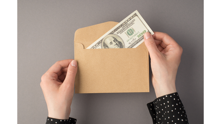 First person top view photo of female hands holding open craft paper envelope with hundred dollars banknote on isolated grey background with copyspace