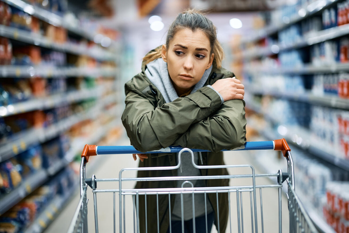 Young sad woman leaning on shopping cart while standing among produce aisle at supermarket.