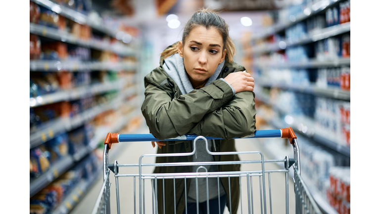 Young sad woman leaning on shopping cart while standing among produce aisle at supermarket.