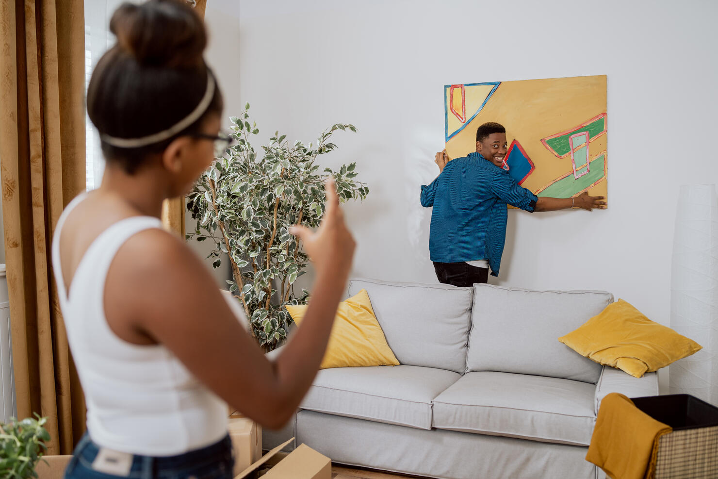 Couple in love decorating their first apartment, unpacking boxes after moving in, the man hanging a painting on the wall, the woman showing how much to lift up to make it even