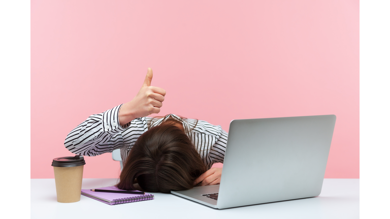 I am fine. Extremely tired sleepy woman lying on table with laptop and cup of coffee showing thumbs up, exhausted with overwork, vitamin deficiency