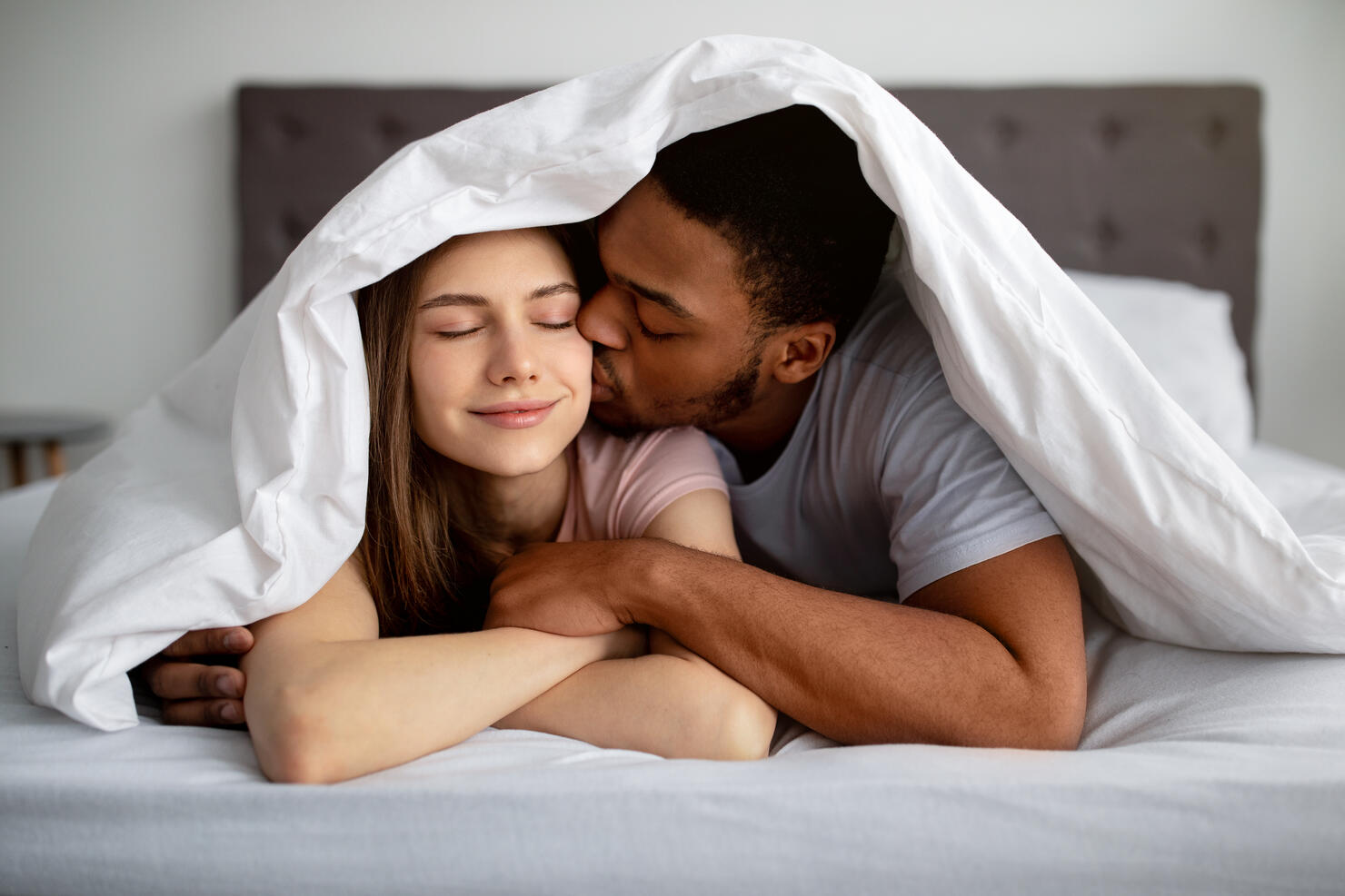 Romantic black guy kissing his Caucasian girlfriend under blanket on bed, indoors