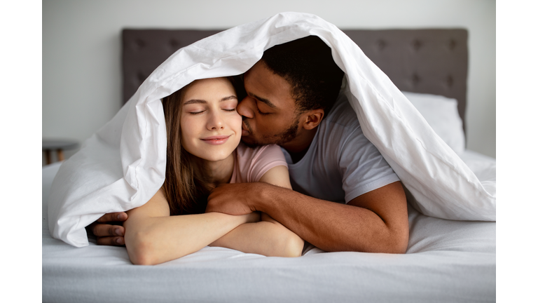 Romantic black guy kissing his Caucasian girlfriend under blanket on bed, indoors