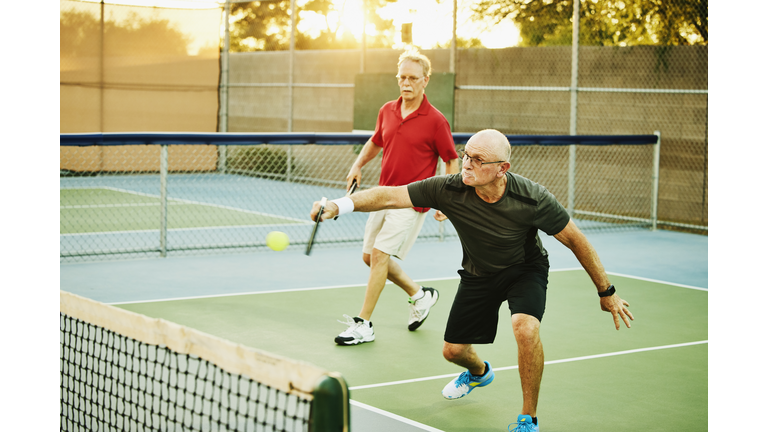 Wide shot of senior man hitting backhand shot at net while playing doubles pickleball on summer evening