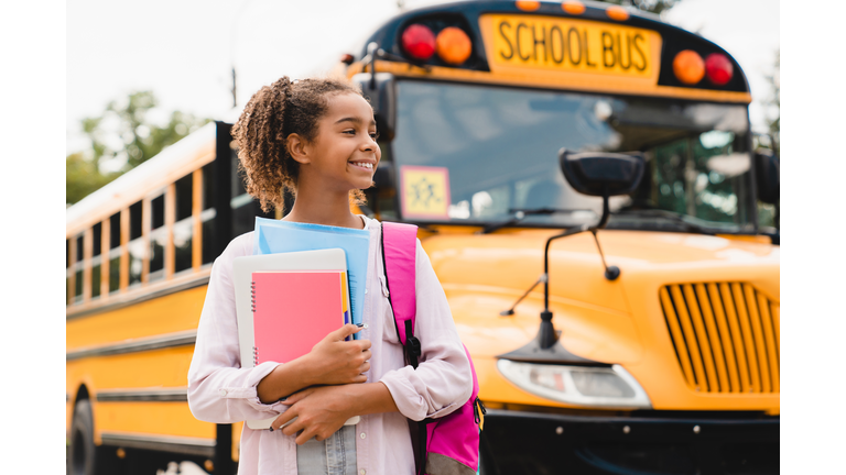 African-american girl teenager pupil student preparing to go to school after summer holidays holding books and notebooks standing next to the school bus.