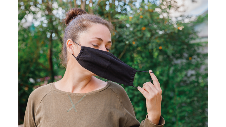 Happy positive girl takes off protective medical mask from face outdoors. Young woman removing mask smiling after vaccination. Coronavirus pandemic Covid 19 concept. Spring flowers pollen allergy