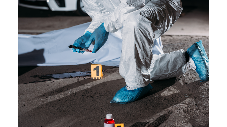 cropped image of criminologist in latex gloves and protective suit holding knife above blood on ground near corpse in body bag at crime scene