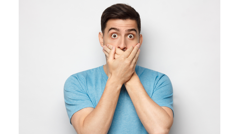 Shocked man dressed in blue t-shirt, covering mouth with both hands, eyes round with shock and fear, isolated on gray background