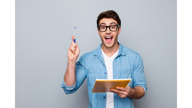 Handsome, attractive, glad, positive, funny guy in glasses with wide open mouth finally found a solution how to make exercise, having raised pen and notebook in hands, isolated on grey background