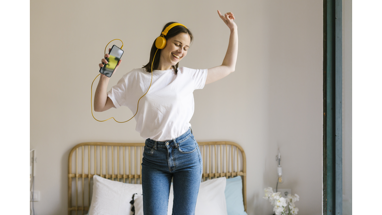 Happy woman with in-ear headphones dancing while holding smart phone in bedroom