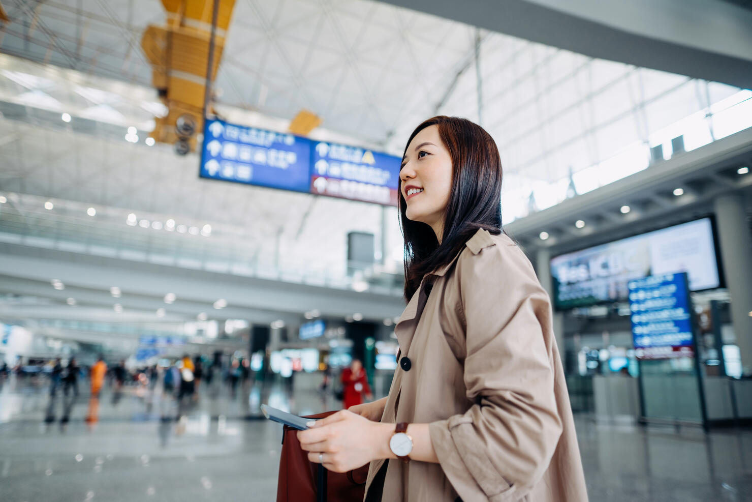 Smiling young Asian woman carrying suitcase and holding passport walking in airport terminal. Ready to travel. Travel and vacation concept. Business person on business trip