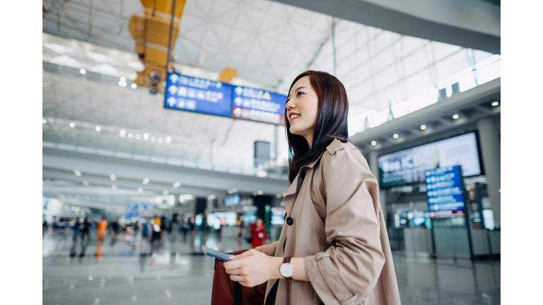 Smiling young Asian woman carrying suitcase and holding passport walking in airport terminal. Ready to travel. Travel and vacation concept. Business person on business trip