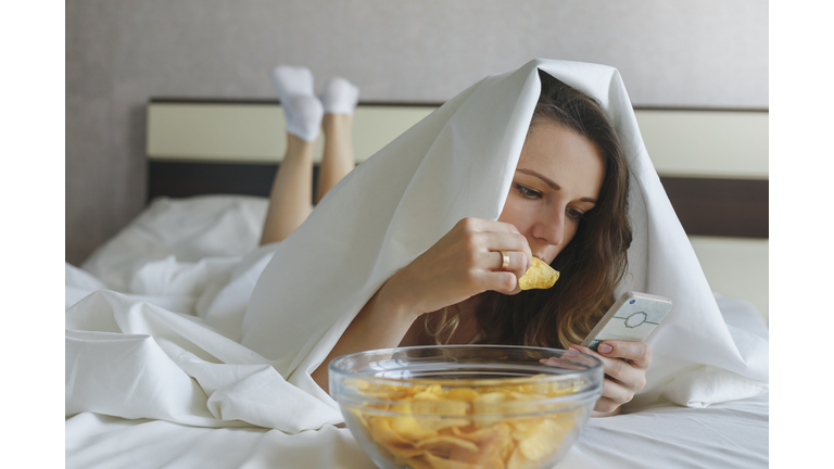 Girl eating chips in bed and watching news in phone
