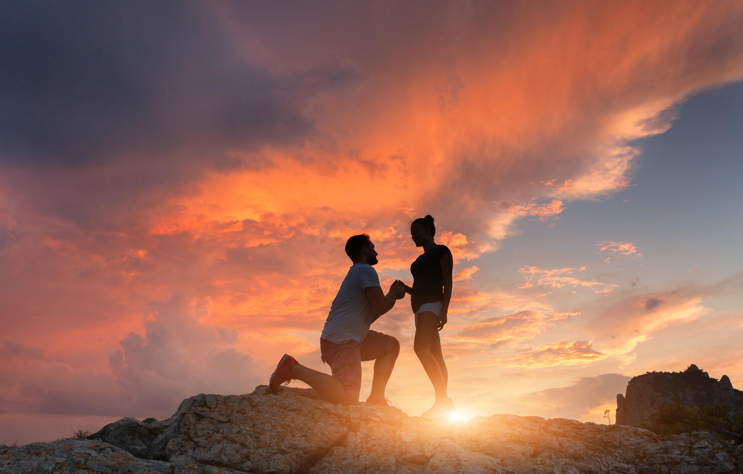 Silhouettes of a man making marriage proposal to his girlfriend on the mountain peak at sunset. Landscape with silhouette of lovers against colorful sky. Couple. People, relationship. Traveling couple