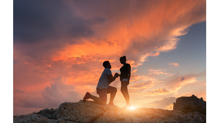 Silhouettes of a man making marriage proposal to his girlfriend on the mountain peak at sunset. Landscape with silhouette of lovers against colorful sky. Couple. People, relationship. Traveling couple