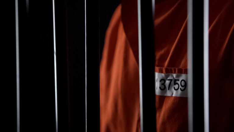 Prisoner in orange uniform standing behind bars, punishment for committed crime