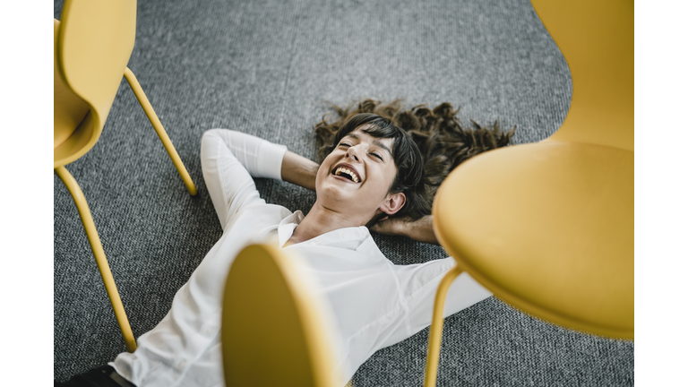 Laughing businesswoman laying in an office on the floor between chairs