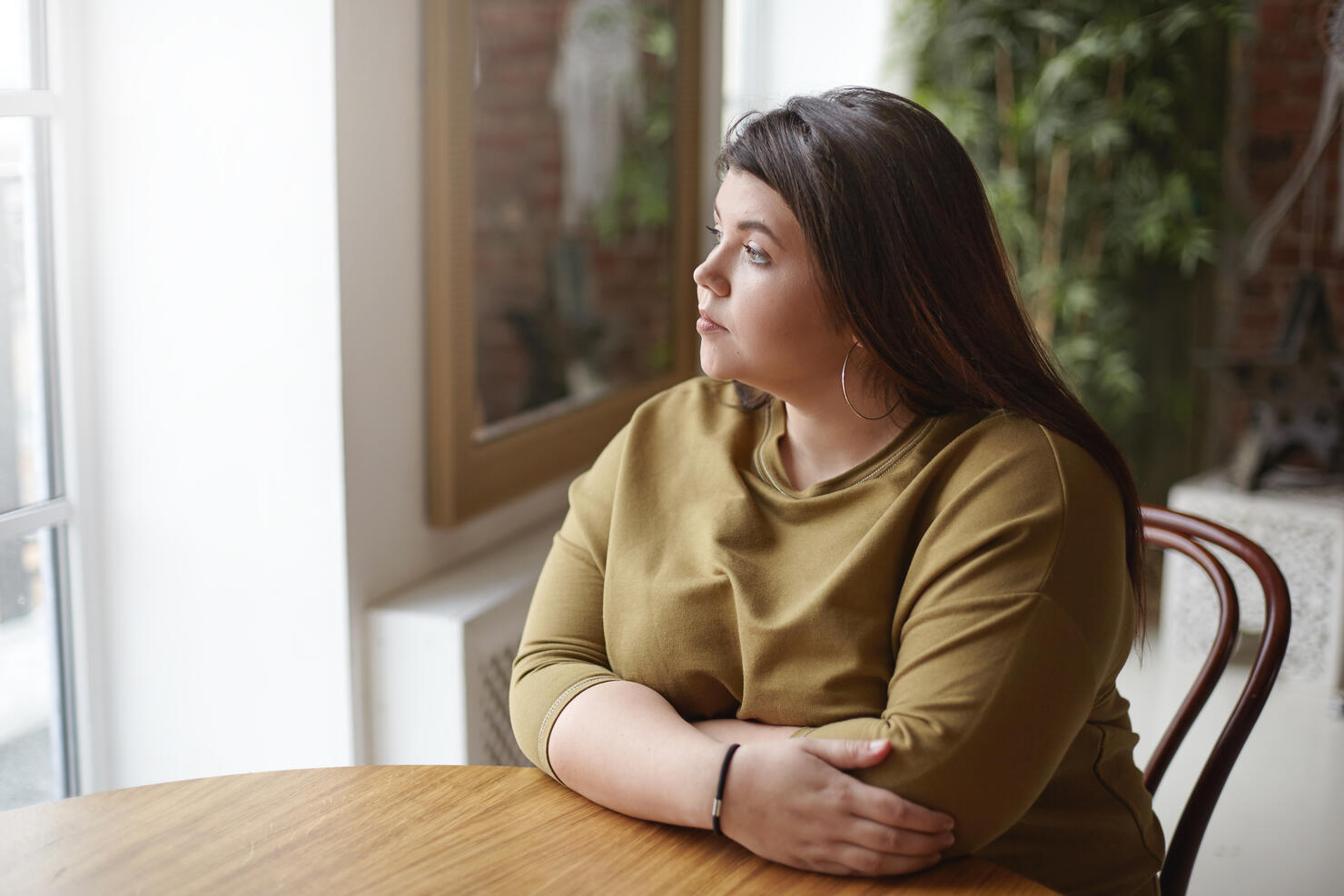 Loneliness concept. Young brunette plus size woman with black hair sitting at cafe table, feeling lonely, spending time alone, waiting for her lunch, looking through window with sad pensive expression