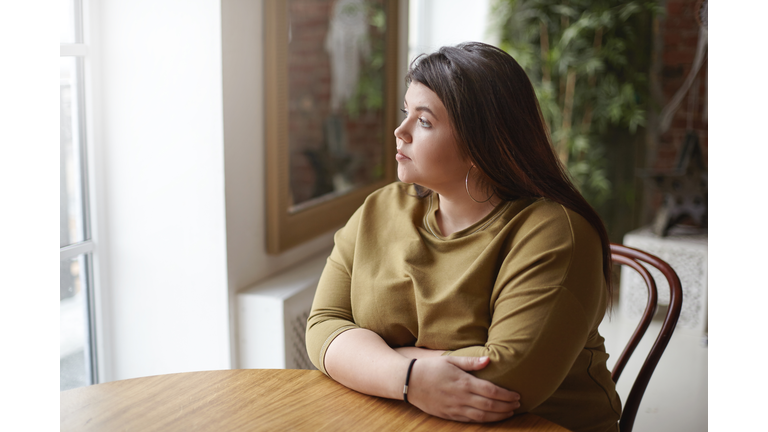 Loneliness concept. Young brunette plus size woman with black hair sitting at cafe table, feeling lonely, spending time alone, waiting for her lunch, looking through window with sad pensive expression