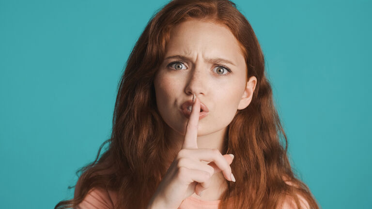 Angry redhead girl seriously showing silence gesture on camera over colorful background. Don't tell expression