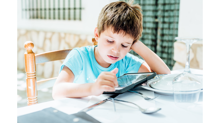 Five years old boy with tablet sitting on a chair and leaning on a table (outdoors)