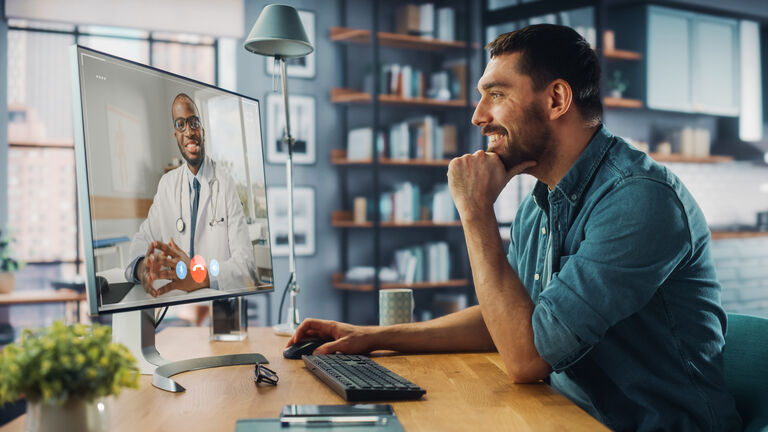 Handsome Caucasian Male is Making a Video Call to His Medical Consultant on Desktop Computer at Home Living Room while Sitting at Table. Man Working From Home and Talking to a Doctor Over the Internet