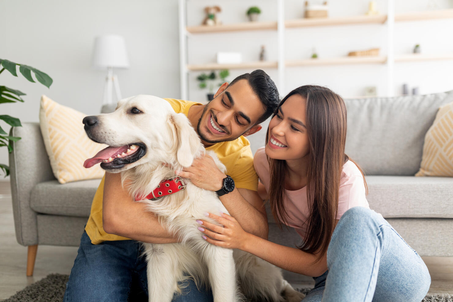 Portrait of happy multiracial couple scratching their pet dog, sitting on floor at home