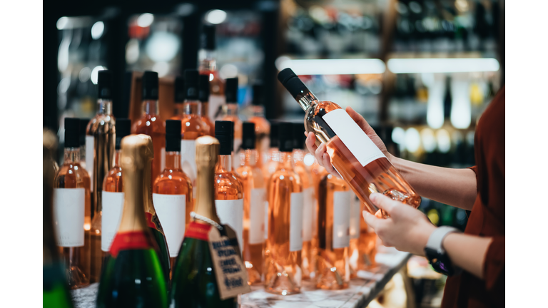 Close up of young Asian woman walking through liquor aisle and choosing a bottle of rose wine from the shelf in supermarket