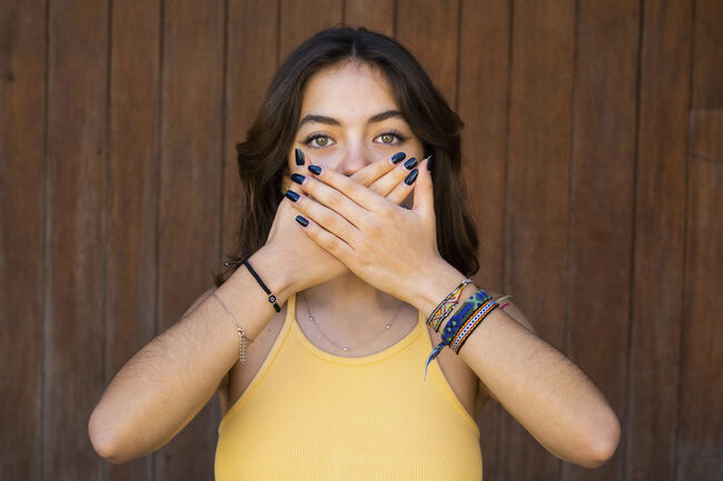 Young Latino woman looking at the camera and covering her mouth, SPEAK NO EVIL, brown wooden background