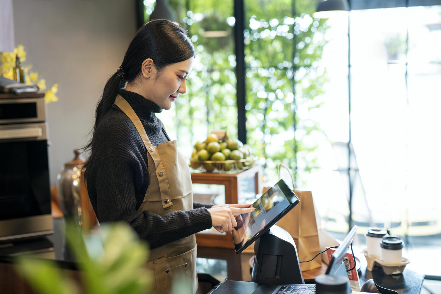 An Asian woman coffee shop owner at the cashier counter is checking and making online orders from customers. Point of Sale System, Take-out Foods, and Small Business Concepts.