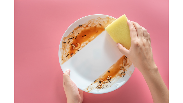 woman using yellow cleaning sponge to clean up and washing food stains and dirt on white dish after eating meal isolated on pink background. cleaning , healthcare and sanitation at home concept