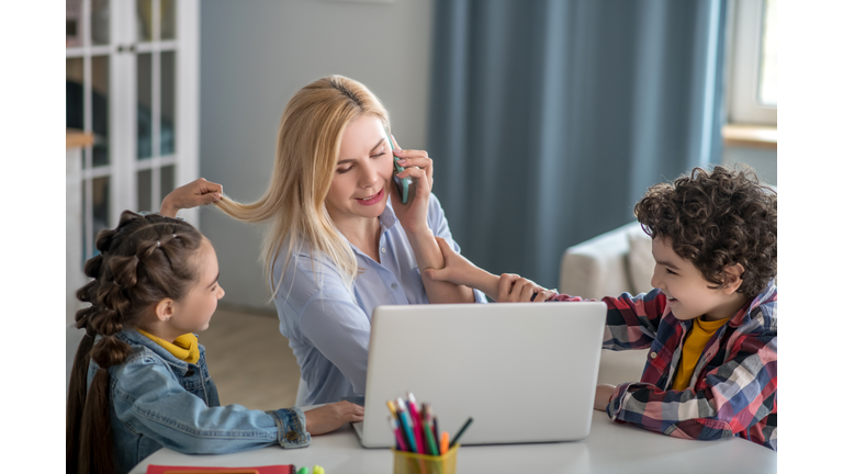 Blonde female talking on mobile and working on laptop, distracted by curly boy and dark-haired girl