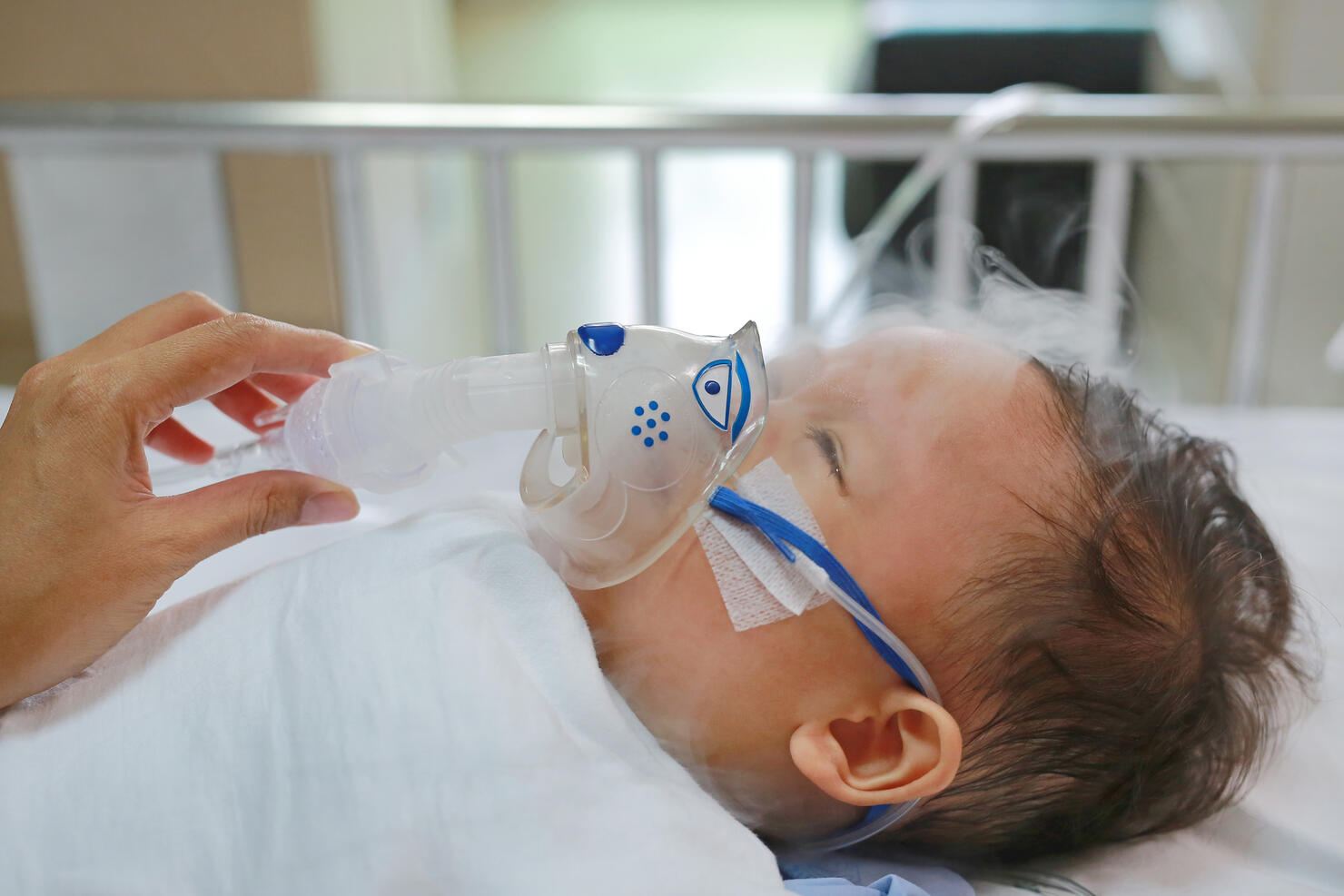 Toddler boy using nebulizer to cure asthma or pneumonia disease . Sick baby boy rest on patients bed and has inhalation therapy by the mask of inhaler. Respiratory Syncytial Virus (RSV).