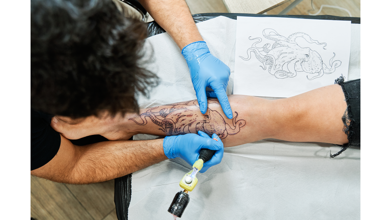 Top view of an unrecognizable tattoo artist tattooing an octopus design on his client's leg.