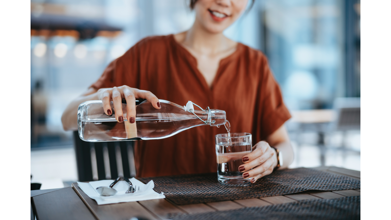 Young Asian woman pouring water from bottle into the glass at outdoor restaurant. Healthy lifestyle and stay hydrated