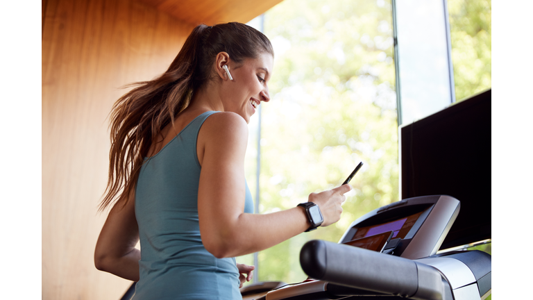 Woman Exercising On Treadmill Wearing Wireless Earphones And Smart Watch Checking Mobile Phone