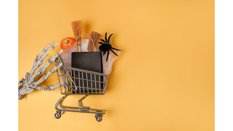 Skeleton hand holding halloween shopping cart on orange background, copy space