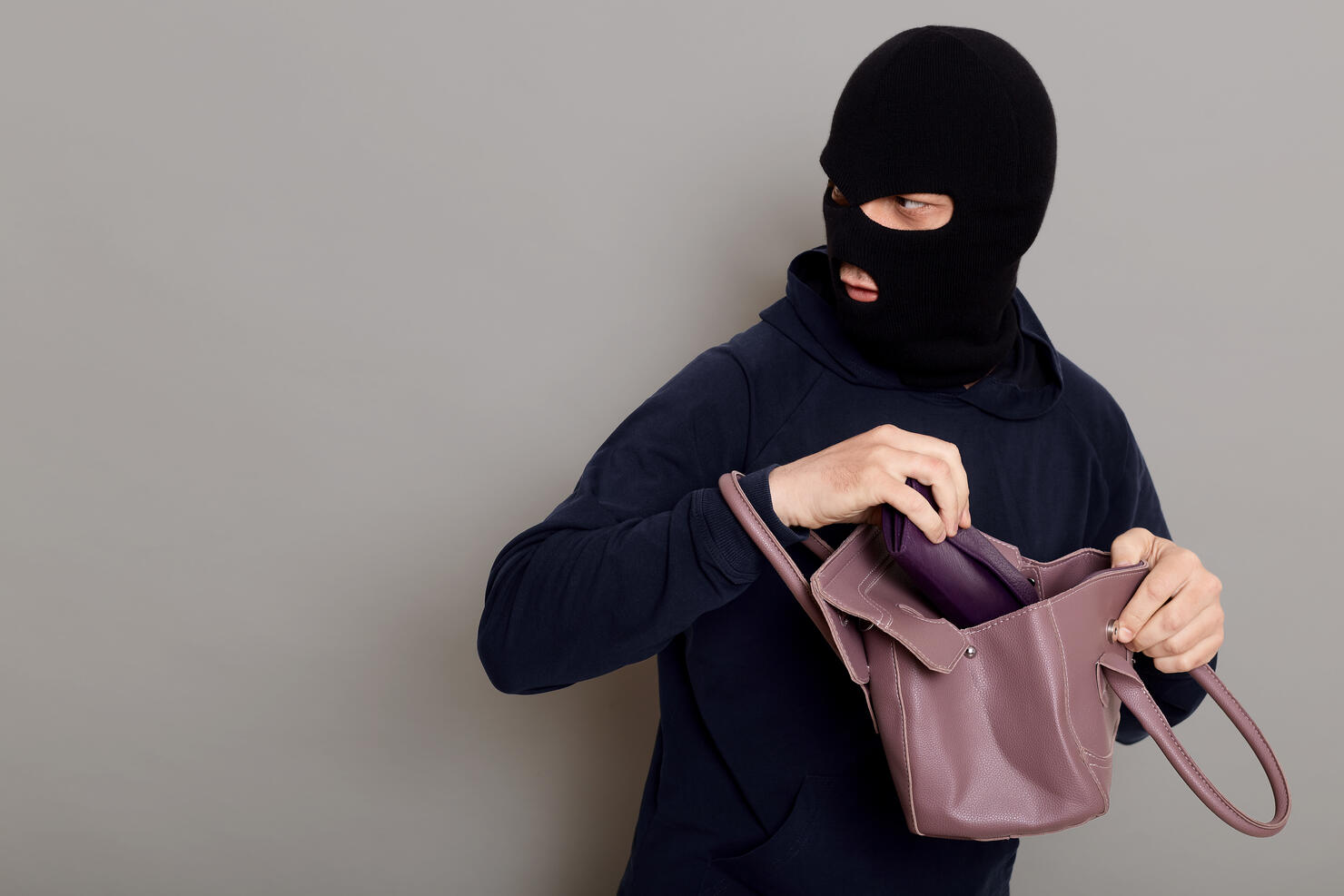 Profile of a thief guy with a masked face, a burglar stole a purse and a woman's handbag looking back, afraid to be caught, copy space, isolated on gray background.