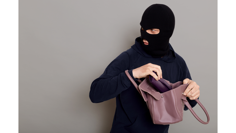 Profile of a thief guy with a masked face, a burglar stole a purse and a woman's handbag looking back, afraid to be caught, copy space, isolated on gray background.