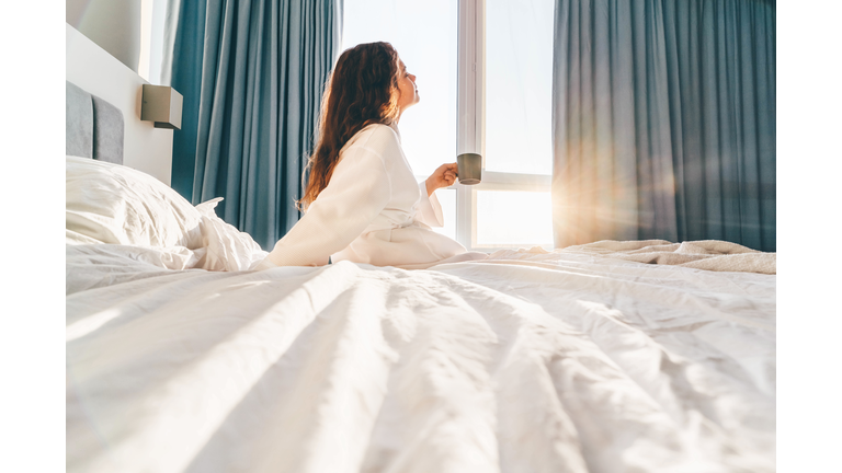 Pretty positive woman with long hair drinks water and sitting at bed in sunny lazy morning.